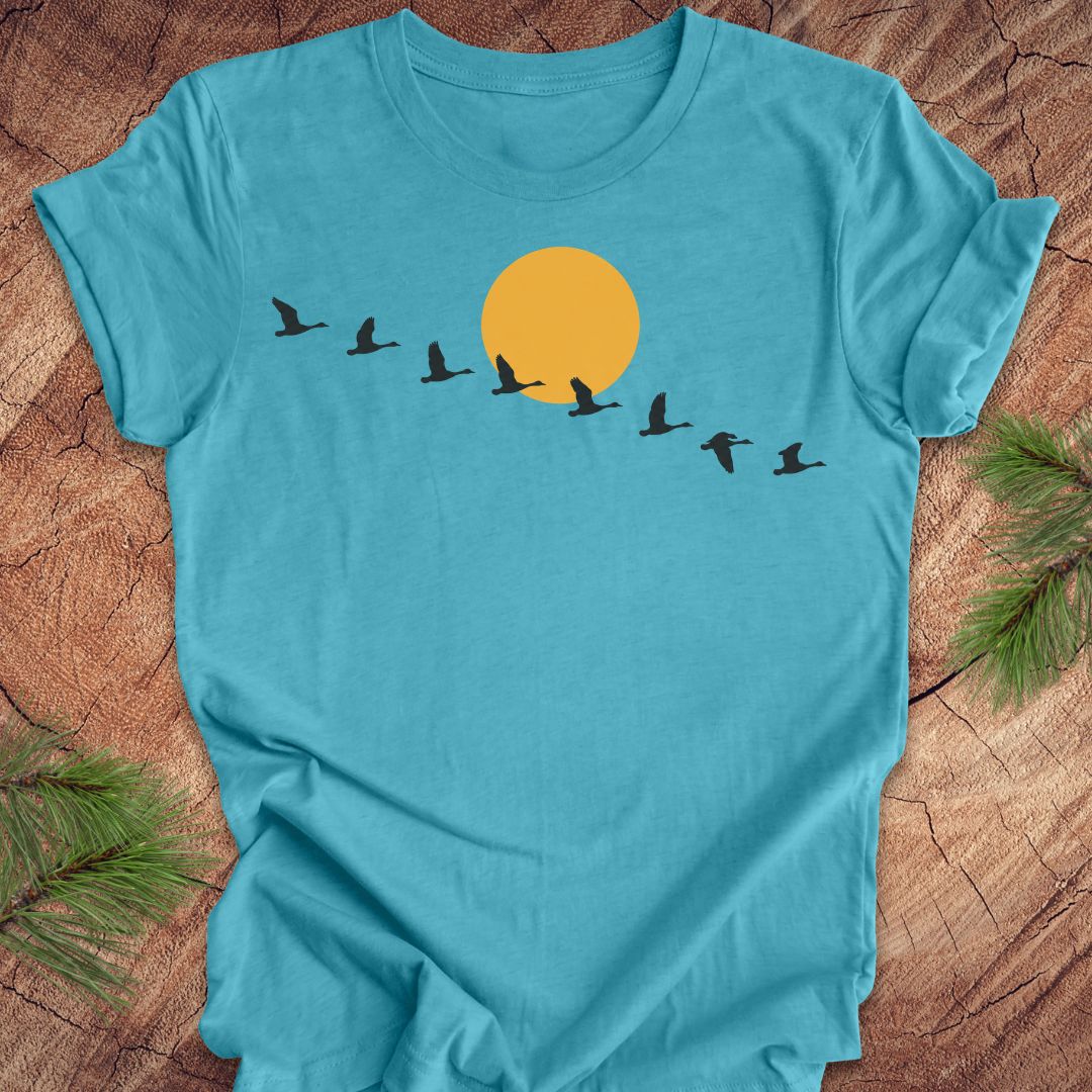 A turquoise blue short-sleeve t-shirt laid flat on a rustic wood surface, featuring a graphic of a large yellow sun and a line of black flying birds across the chest. Green pine branches are placed in the corners as styling props.