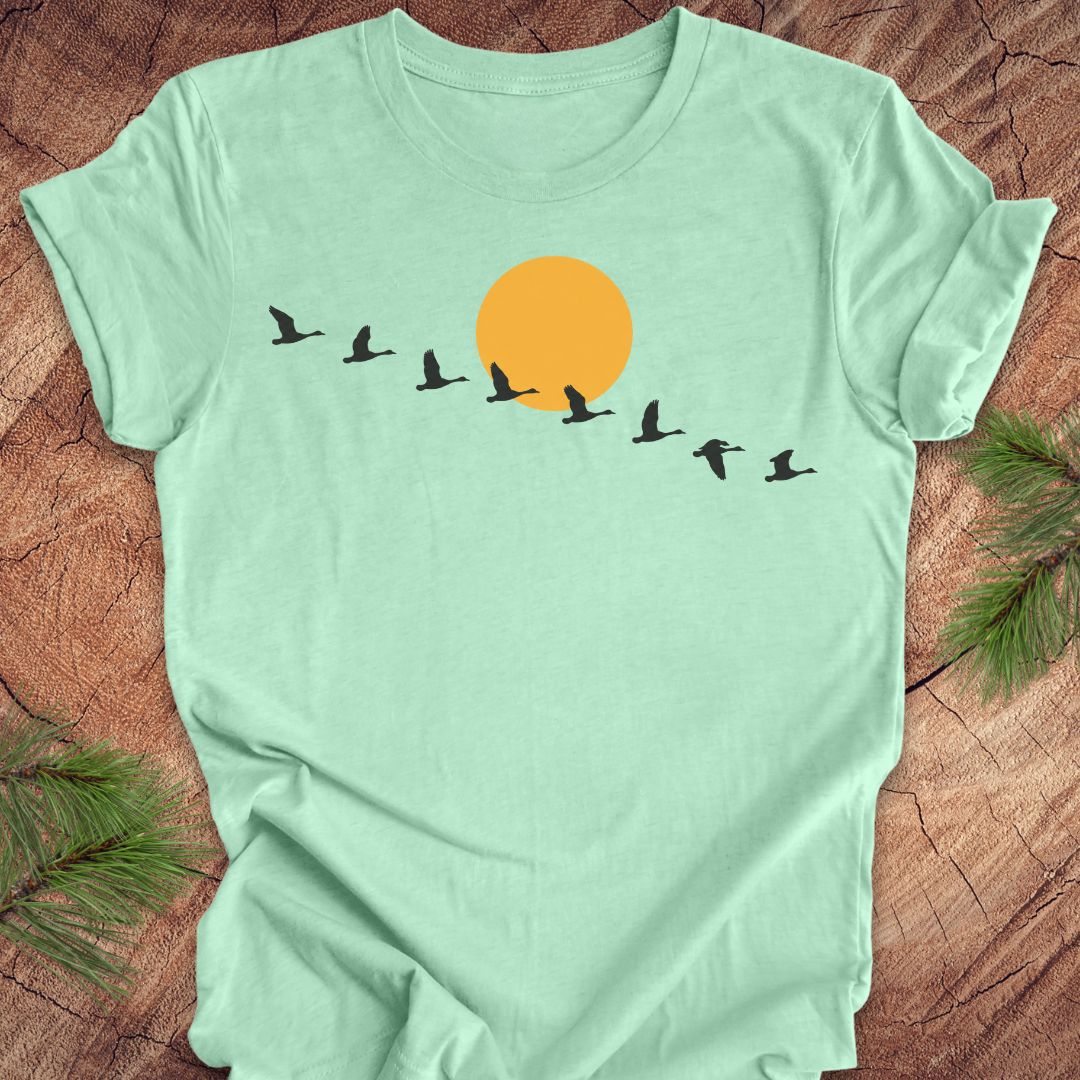 A mint green short-sleeve t-shirt laid flat on a rustic wood surface, featuring a graphic of a large orange sun and a line of black flying birds across the chest. Pine branches are visible in the corners.
