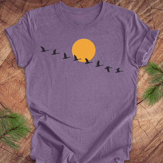 A purple short-sleeve t-shirt laid flat on a rustic wood surface, featuring a graphic of a large orange sun with black bird silhouettes flying in front. Green pine branches are placed in the corners of the frame.