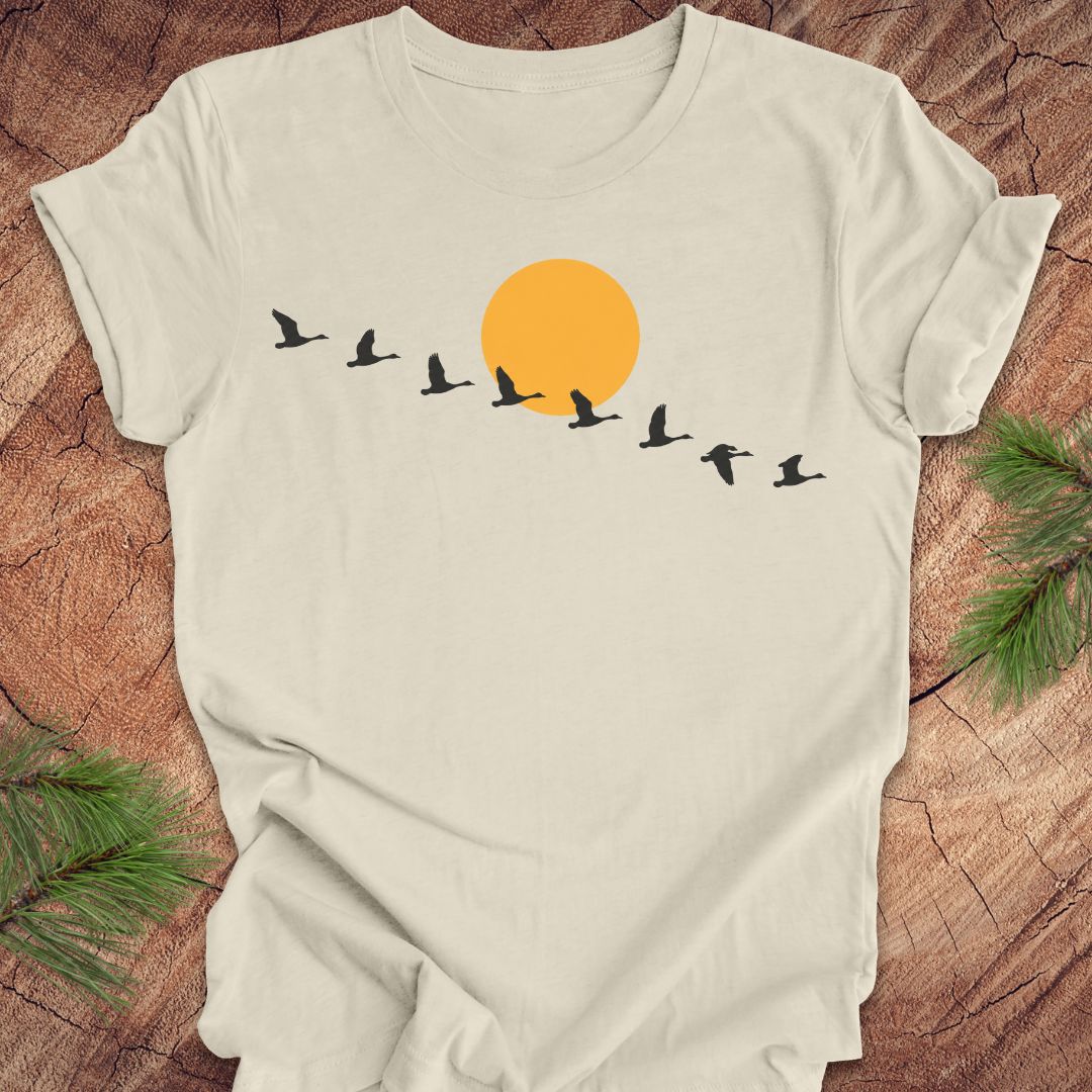 An off-white short-sleeve t-shirt laid flat on a rustic wood background, featuring a graphic of a large orange sun and black flying birds across the chest. Green pine branches are visible in the corners.