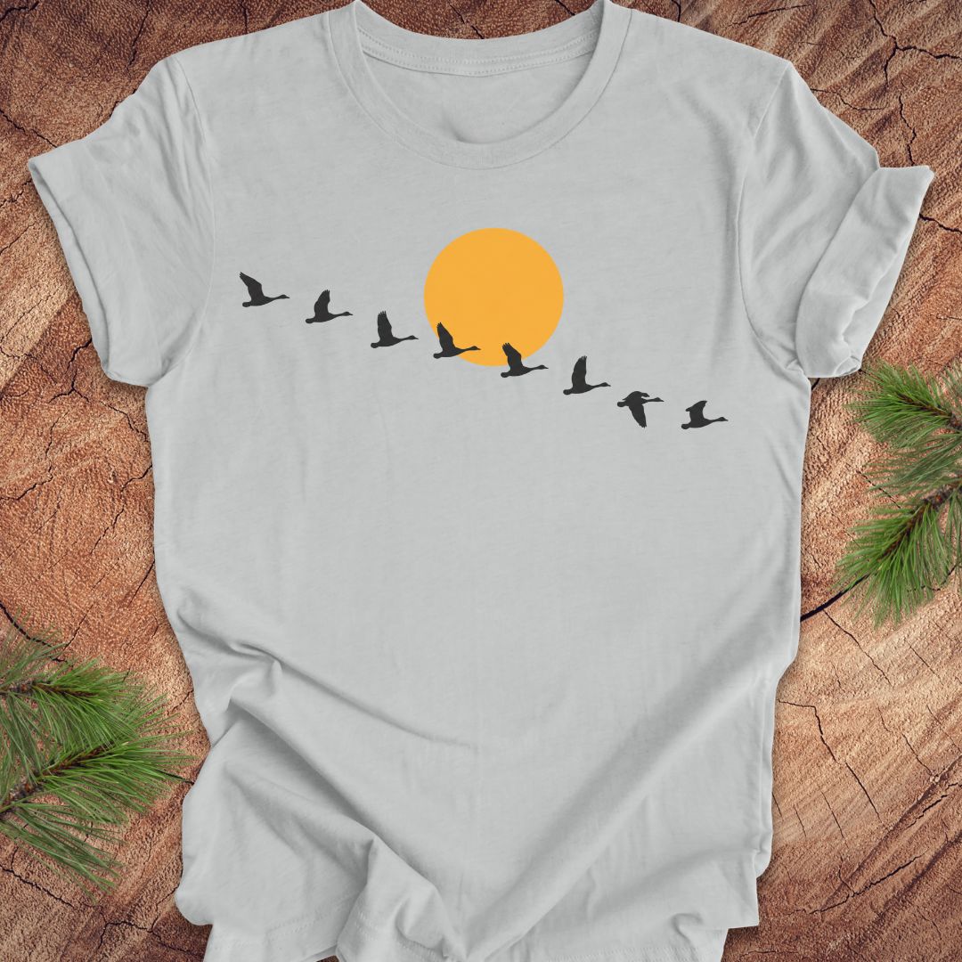 A light gray short-sleeve t-shirt laid flat on a rustic wood surface, featuring a graphic of a large orange sun and black flying birds. Green pine branches are placed in the corners of the scene.