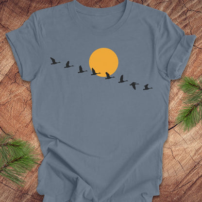 A slate blue crewneck t-shirt laid flat on a rustic wood background with pine branches in the corners. The shirt features a graphic of a large orange sun with a line of black flying birds in front of it.