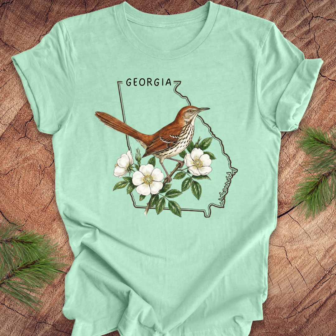 Mint green t-shirt with the outline of the state of Georgia and state bird and flower, brown thrasher and cherokee rose on a wooden surface