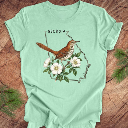 Mint green t-shirt with the outline of the state of Georgia and state bird and flower, brown thrasher and cherokee rose on a wooden surface