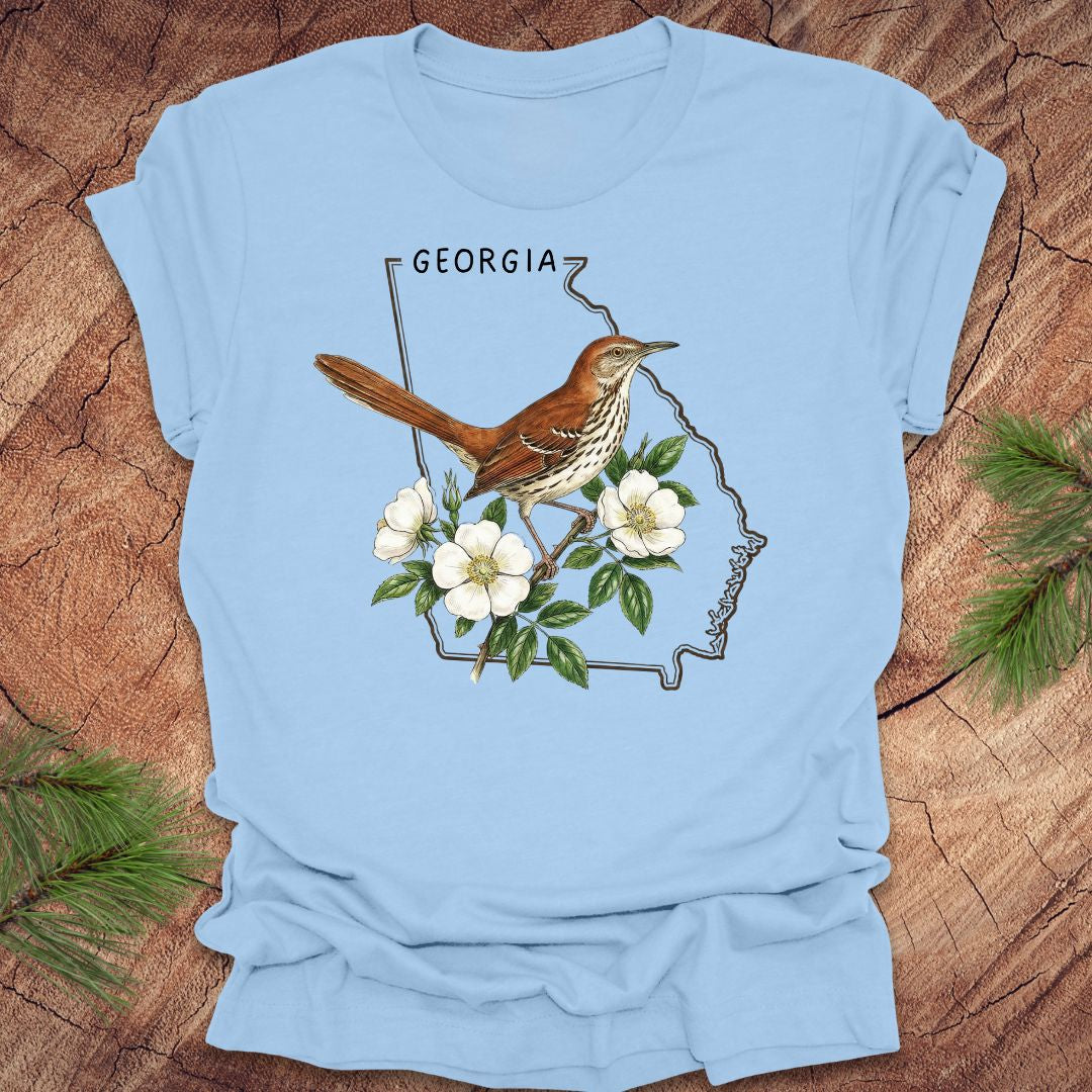 Baby blue t-shirt with the outline of the state of Georgia and state bird and flower, brown thrasher and cherokee rose on a wooden surface