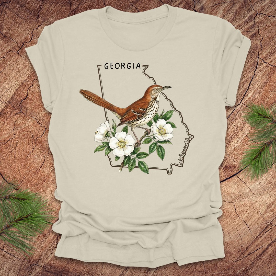 Beige t-shirt with the outline of the state of Georgia and state bird and flower, brown thrasher and cherokee rose on a wooden surface