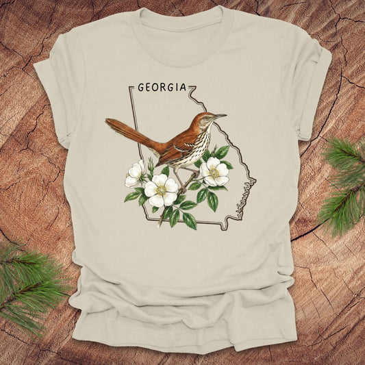 Beige t-shirt with the outline of the state of Georgia and state bird and flower, brown thrasher and cherokee rose on a wooden surface