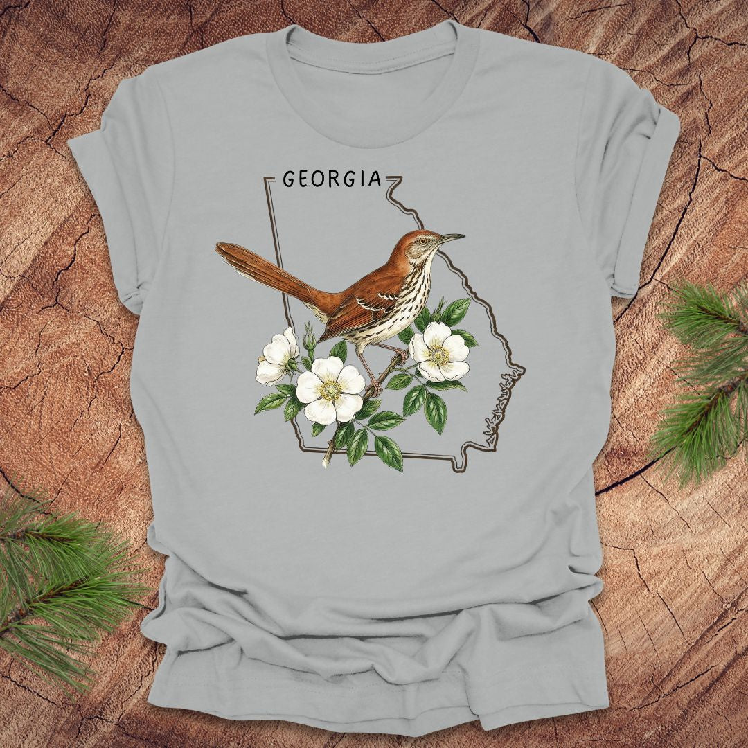 Silver t-shirt with the outline of the state of Georgia and state bird and flower, brown thrasher and cherokee rose on a wooden surface