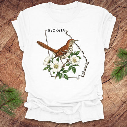 White t-shirt with the outline of the state of Georgia and state bird and flower, brown thrasher and cherokee rose on a wooden surface