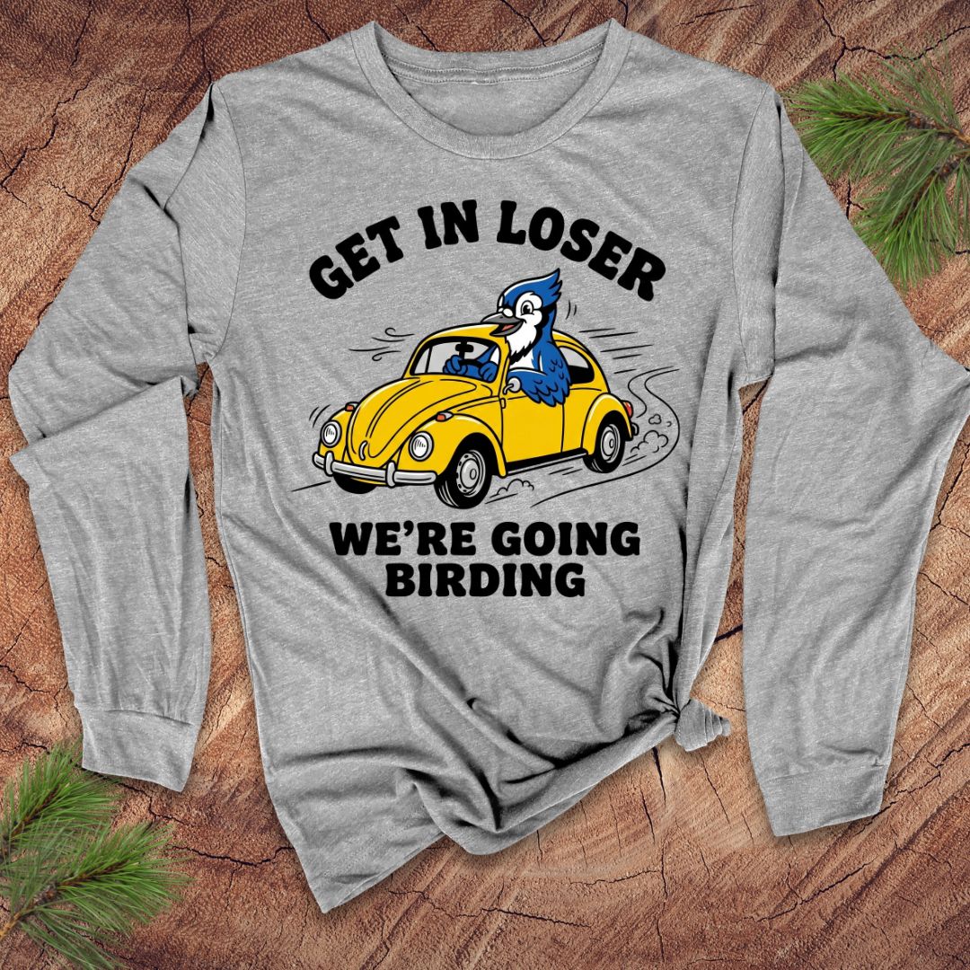 Gray long-sleeve shirt with a graphic of a yellow car and blue bird, text 'GET IN LOSER WE'RE GOING BIRDING', on a wooden surface.