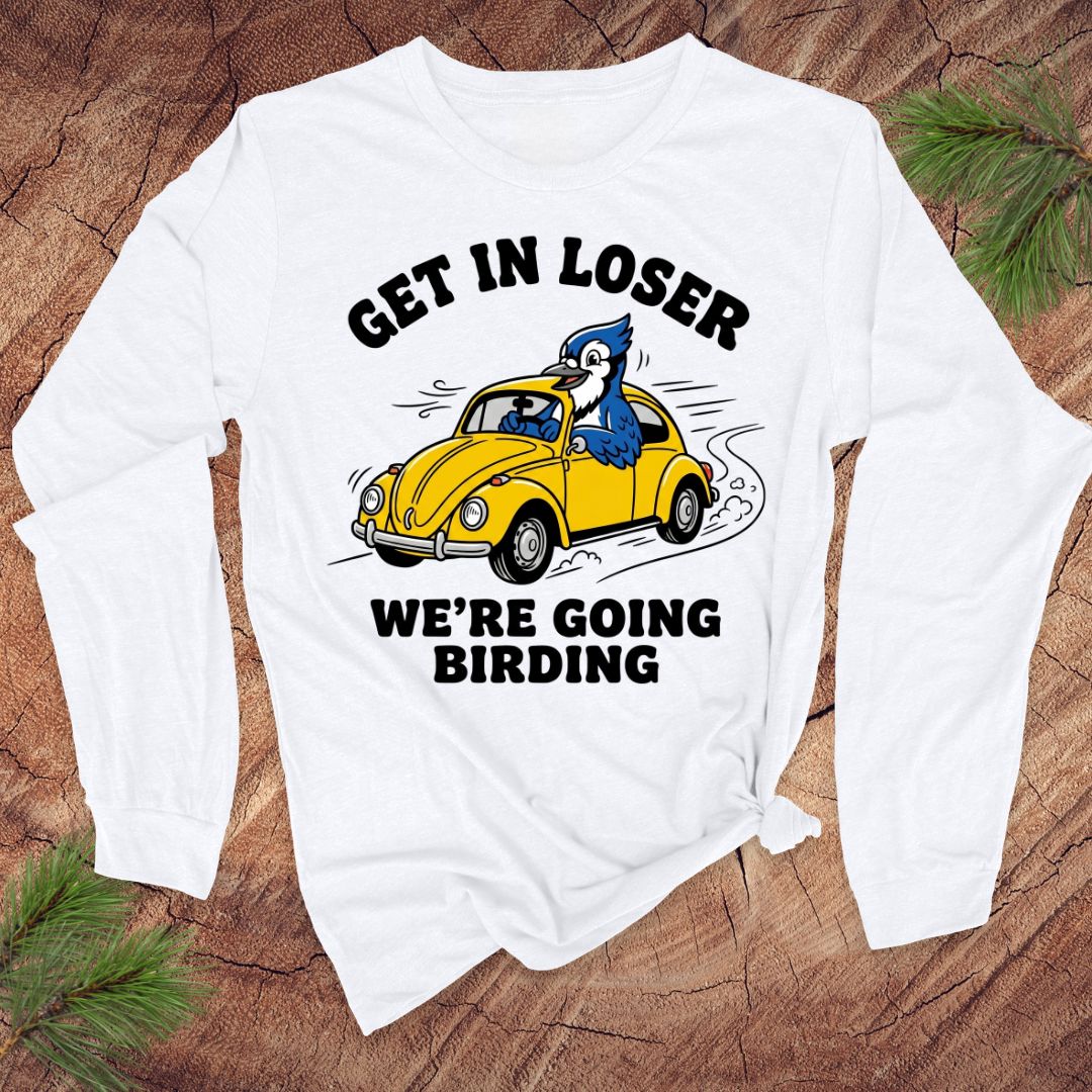 White long-sleeve shirt with a graphic of a yellow car and blue bird, text 'GET IN LOSER WE'RE GOING BIRDING' on a wooden surface.