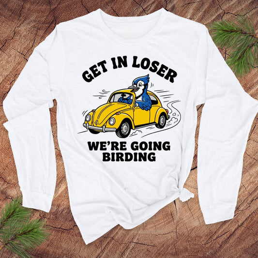 White long-sleeve shirt with a graphic of a yellow car and blue bird, text 'GET IN LOSER WE'RE GOING BIRDING' on a wooden surface.