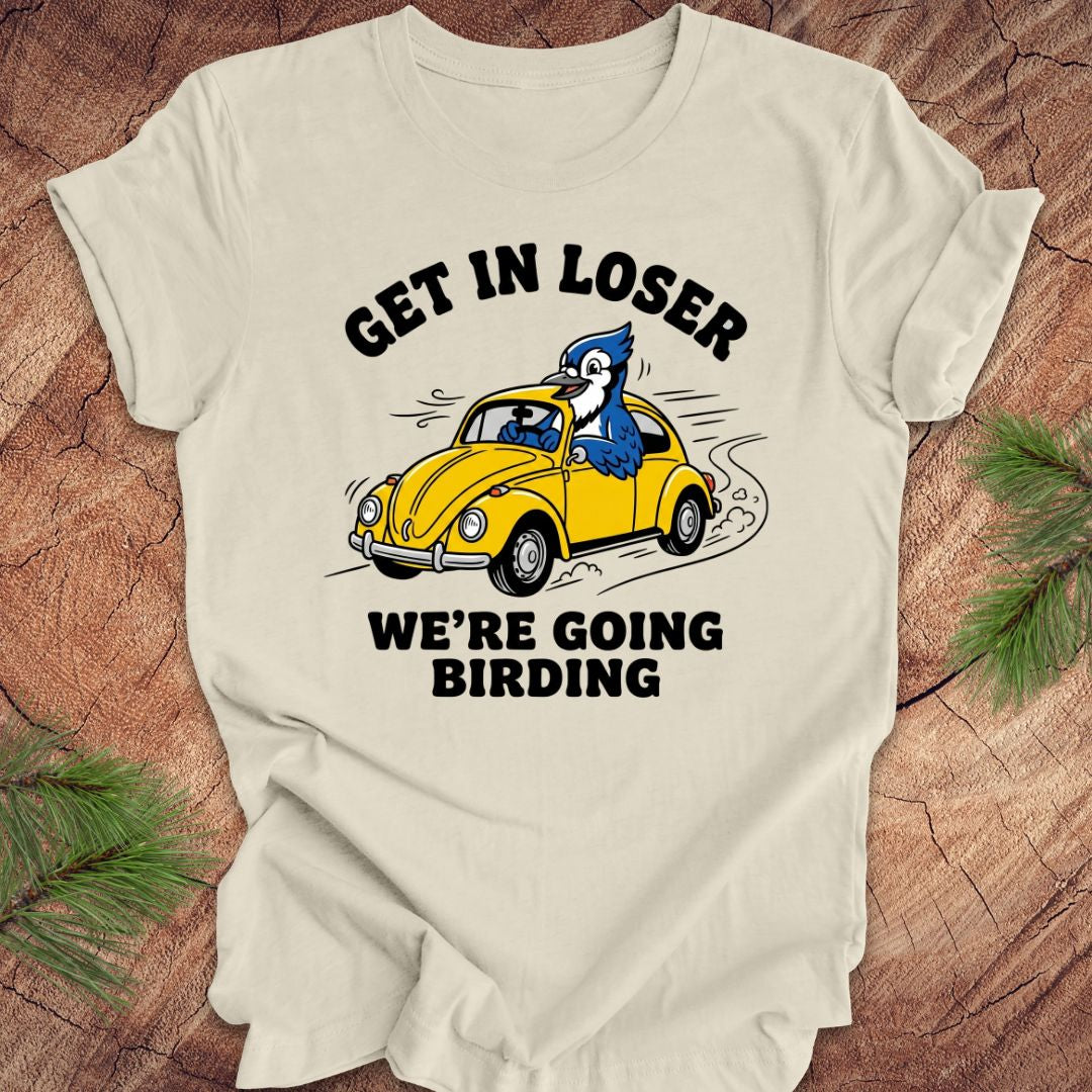 Beige t-shirt with a graphic of a blue jay driving a yellow car and text on a wooden surface.