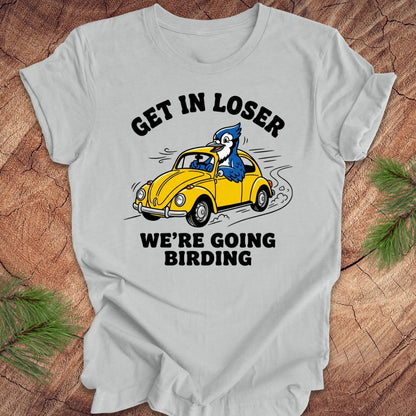 Silver t-shirt with a graphic of a blue jay driving a yellow car and text on a wooden surface.