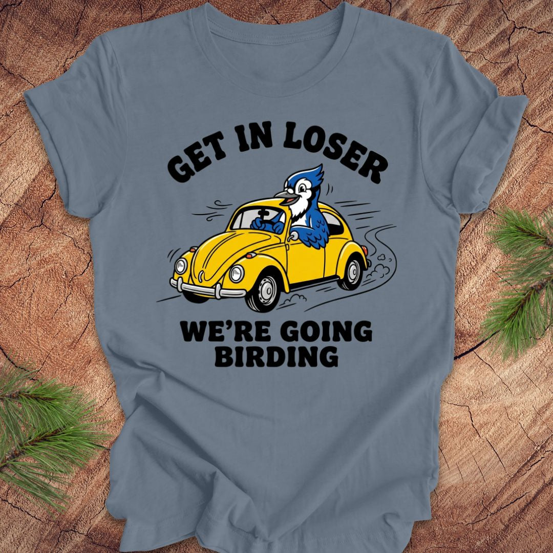 Blue t-shirt with a graphic of a yellow car and blue jay, text 'GET IN LOSER WE'RE GOING BIRDING', on a wooden surface.