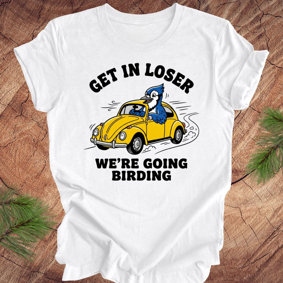 White t-shirt with a graphic of a yellow car and blue jay, text 'GET IN LOSER WE'RE GOING BIRDING' on a wooden background.