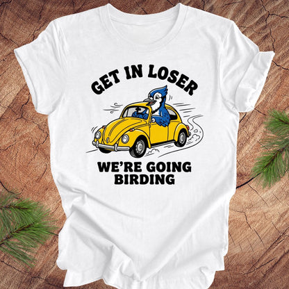 White t-shirt with a graphic of a yellow car and blue jay, text 'GET IN LOSER WE'RE GOING BIRDING' on a wooden background.