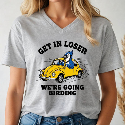 Person wearing a gray v-neck t-shirt with a graphic blue jay design and text, 'get in loser. we're going birding' on a white background.