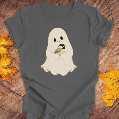 A dark gray short-sleeve t-shirt with a cute cartoon graphic of a smiling ghost holding a small chickadee bird. The t-shirt is laid flat on a rustic wood background decorated with autumn orange and yellow leaves.