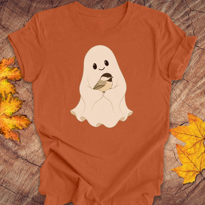 An orange short-sleeve t-shirt laid flat on a rustic wood background, featuring a cute cartoon graphic of a smiling ghost holding a small bird. Yellow and orange autumn leaves are placed on either side of the shirt.