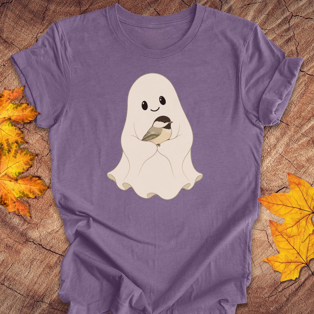 A purple short-sleeve t-shirt laid flat on a rustic wood background, decorated with autumn leaves. The shirt features a cute cartoon graphic of a smiling white ghost holding a small black and white chickadee bird.