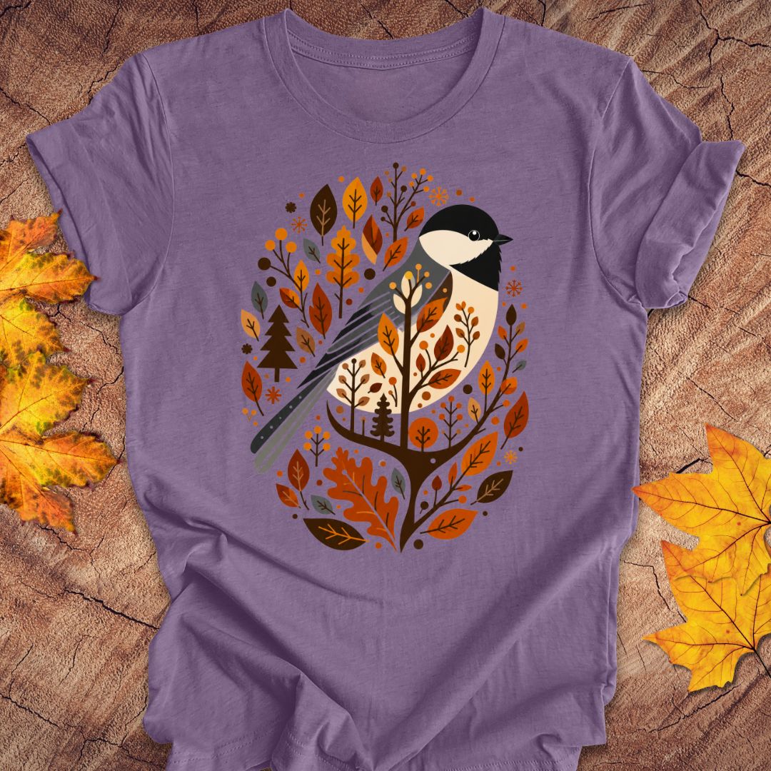 A purple short-sleeve t-shirt laid flat on a rustic wood surface, decorated with autumn leaves. The center of the shirt features a graphic of a bird perched among orange, brown, and beige autumn leaves and trees.