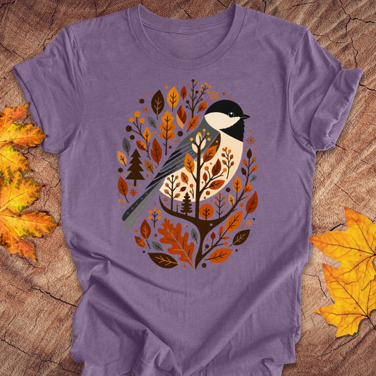A purple short-sleeve t-shirt laid flat on a rustic wood surface, decorated with autumn leaves. The center of the shirt features a graphic of a bird perched among orange, brown, and beige autumn leaves and trees.