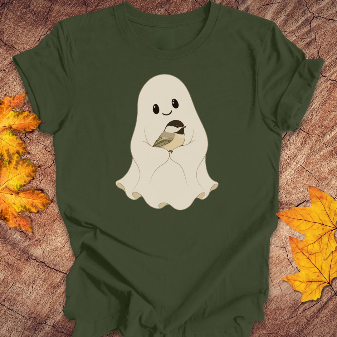 An olive green short-sleeve t-shirt laid flat on a textured wood background, decorated with autumn maple leaves. The shirt features a cute graphic of a smiling cream-colored ghost holding a small chickadee bird.