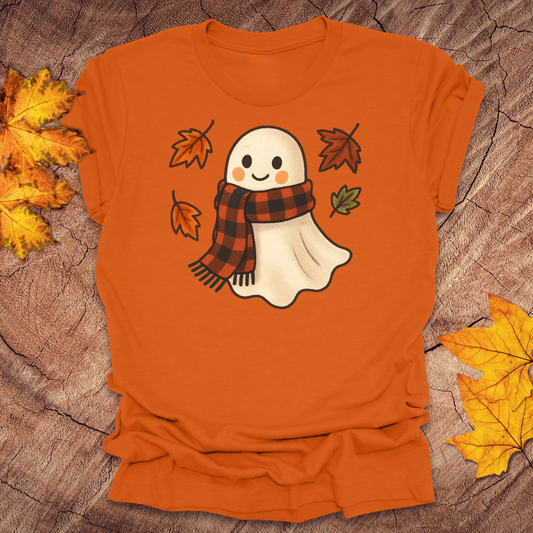 Orange t-shirt with a ghost wearing a plaid scarf design on a wooden surface with autumn leaves.