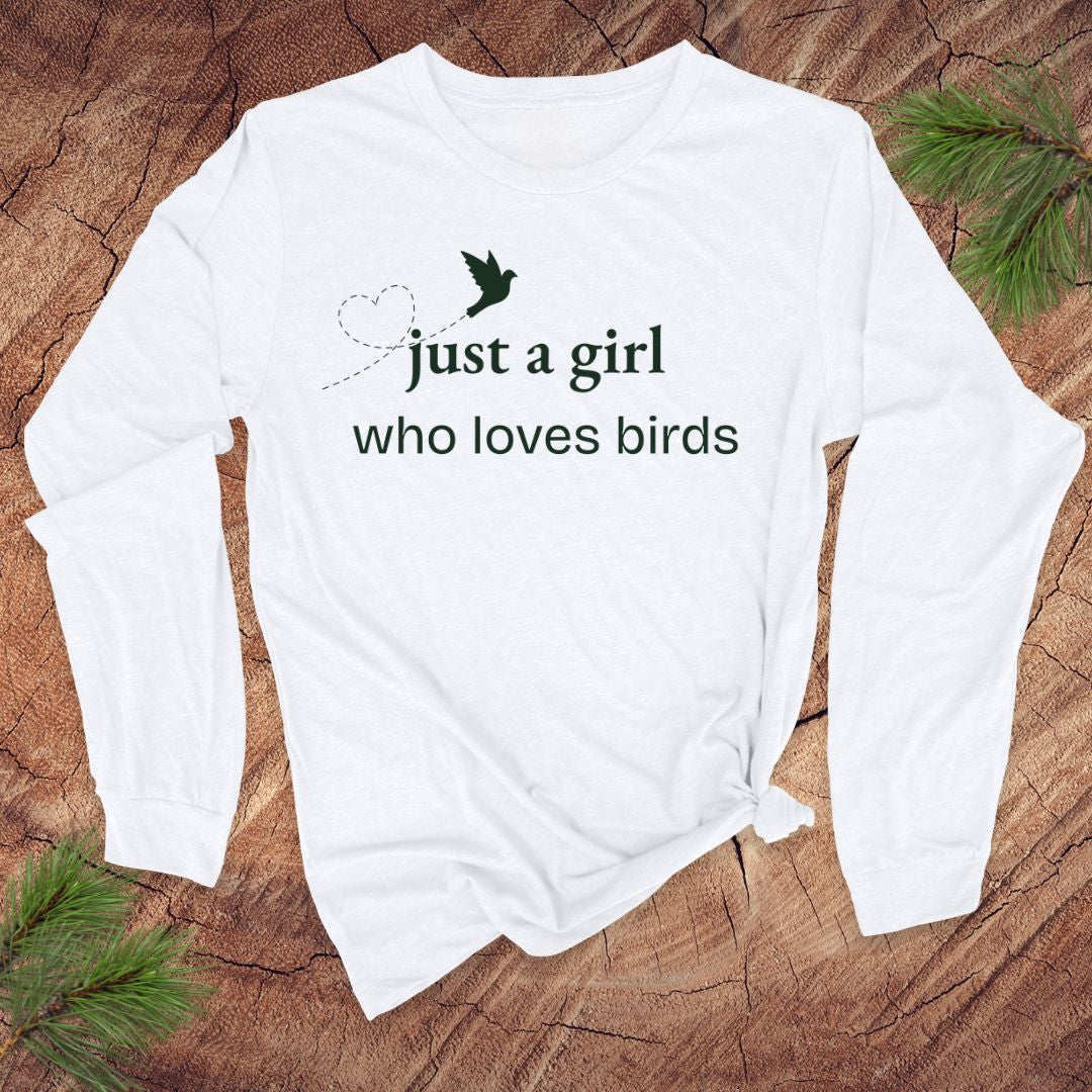White long-sleeve shirt with 'just a girl who loves birds' text on a wooden surface with greenery.
