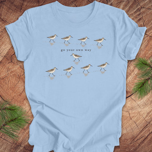 A light blue short-sleeve t-shirt laid flat on a rustic wood background, decorated with green pine branches in the corners. The shirt features a graphic of walking sandpipers with the text 'go your own way' printed in the center.