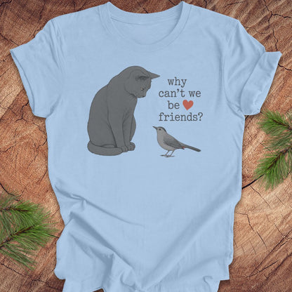 A light blue short-sleeve t-shirt laid flat on a rustic wood surface, featuring a printed design of a gray cat and a small bird with the text 'why can't we be friends?' and a small red heart.