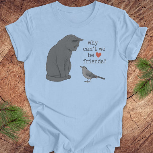 A light blue short-sleeve t-shirt laid flat on a rustic wood surface, featuring a printed design of a gray cat and a small bird with the text 'why can't we be friends?' and a small red heart.