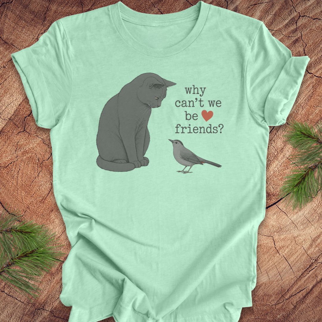 A mint green short-sleeve t-shirt laid flat on a rustic wood surface, featuring a printed design of a gray cat looking at a small gray bird with the text 'why can't we be friends?' and a small red heart.