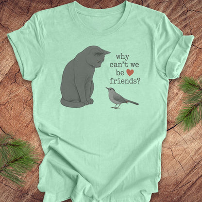 A mint green short-sleeve t-shirt laid flat on a rustic wood surface, featuring a printed design of a gray cat looking at a small gray bird with the text 'why can't we be friends?' and a small red heart.