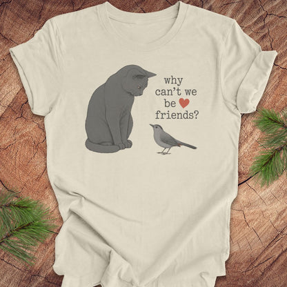 An off-white t-shirt laid flat on a rustic wood background with pine branches. The shirt features a graphic of a gray cat looking at a small gray bird, with the text 'why can't we be friends?' and a small red heart.
