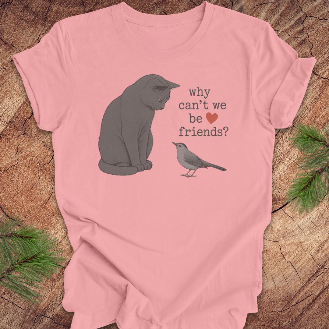 A pink short-sleeve graphic T-shirt laid flat on a rustic wood surface with pine branches around it. The shirt features a gray cat, a small gray bird, and the text 'why can't we be friends?' with a red heart.