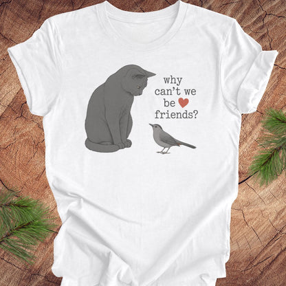 A white short-sleeve crewneck t-shirt with a printed graphic of a gray cat and a small gray bird facing each other, with the text 'why can't we be friends?' and a red heart. The t-shirt is laid flat on a rustic wood background with pine branches in the corners.
