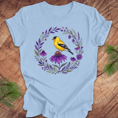 Light blue t-shirt with a goldfinch and purple flowers design on a wooden surface.
