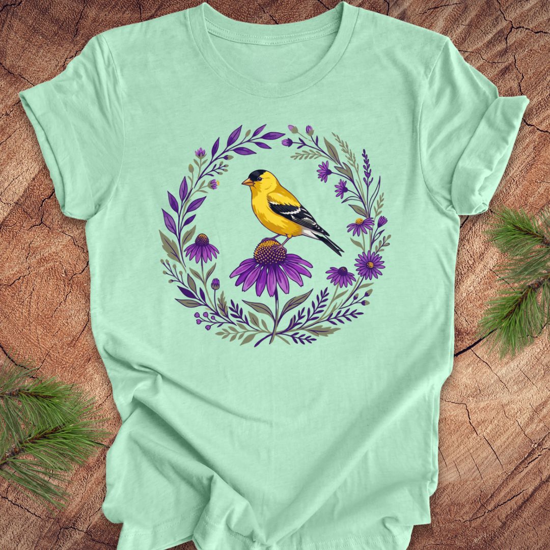 Mint green t-shirt with a goldfinch and purple flowers design on a wooden surface.