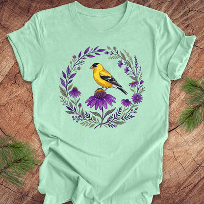 Mint green t-shirt with a goldfinch and purple flowers design on a wooden surface.