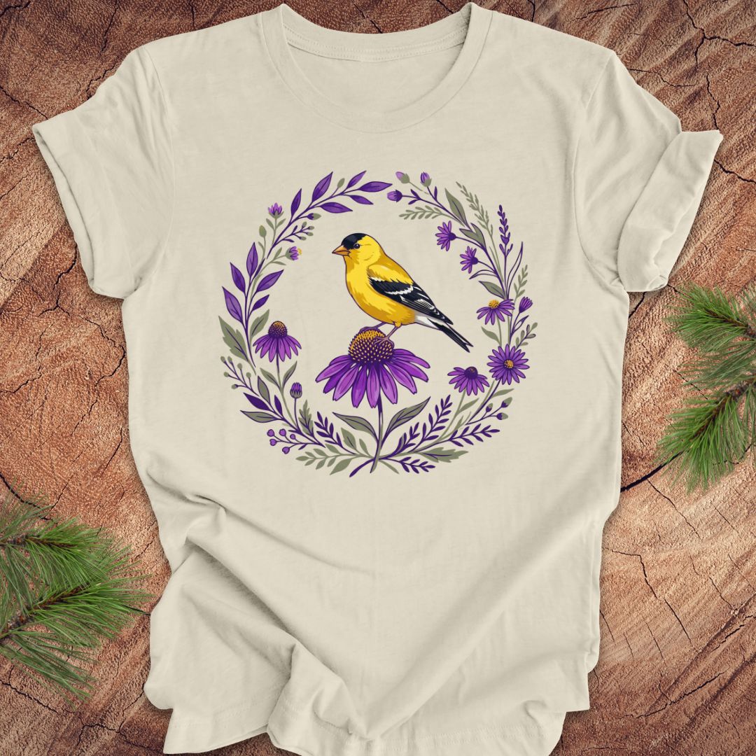Beige t-shirt with a goldfinch and purple flowers design on a wooden surface.