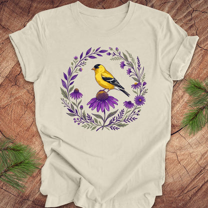 Beige t-shirt with a goldfinch and purple flowers design on a wooden surface.