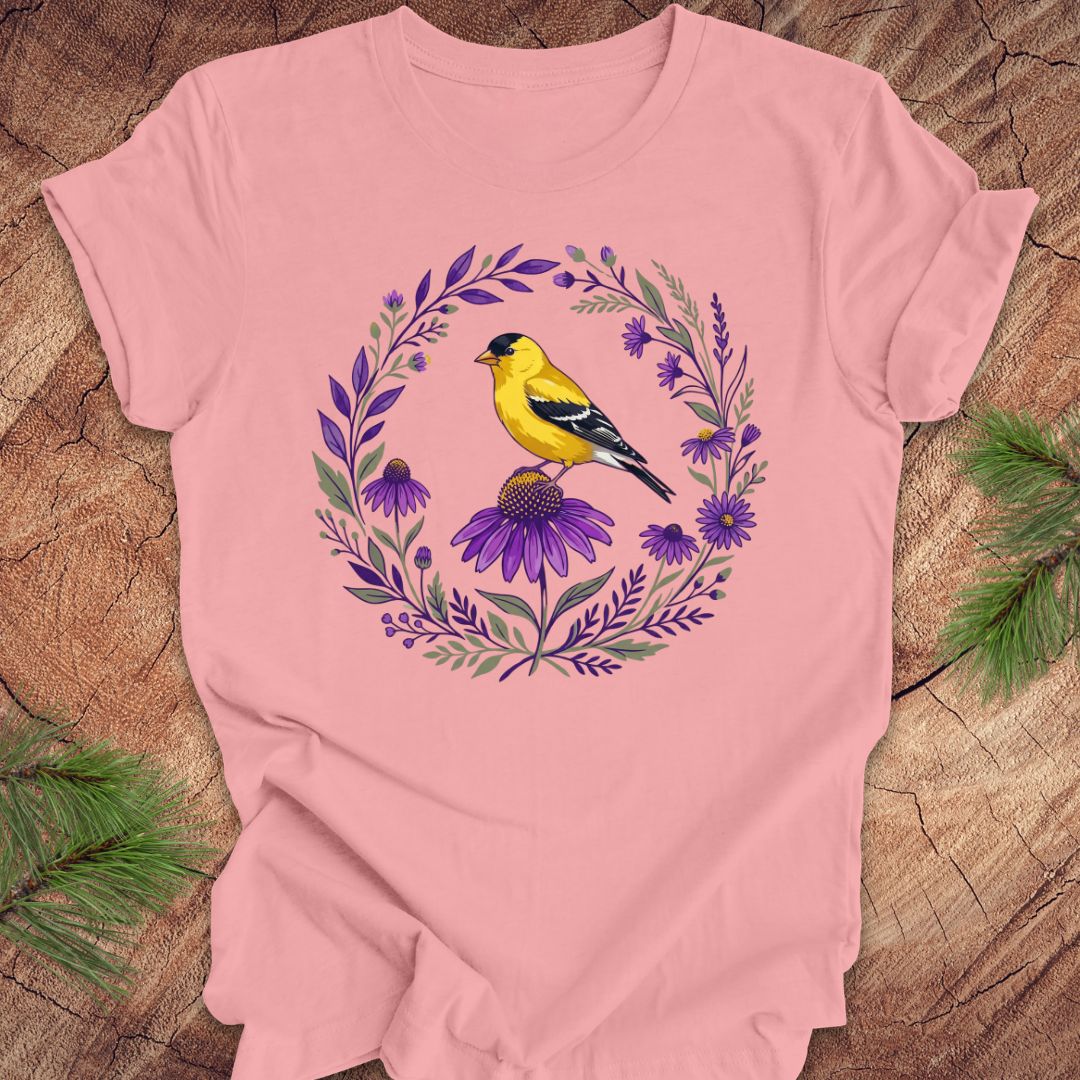 Pink t-shirt with a goldfinch and purple flowers design on a wooden surface.