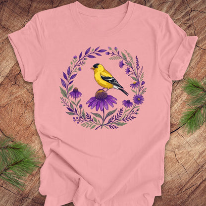 Pink t-shirt with a goldfinch and purple flowers design on a wooden surface.