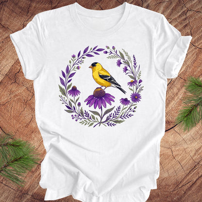 White t-shirt with a goldfinch and purple flowers design on a wooden surface.