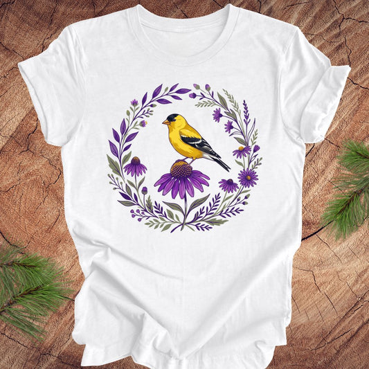 White t-shirt with a goldfinch and purple flowers design on a wooden surface.