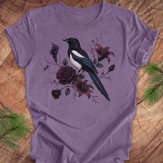 A purple short-sleeve t-shirt with a graphic of a magpie bird perched among dark red and purple roses, laid flat on a rustic wood background with pine branches in the corners.
