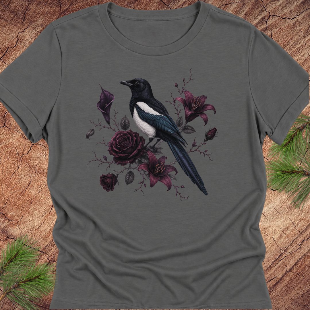 A dark gray short-sleeve crewneck t-shirt featuring a graphic print of a black and white magpie perched among dark purple roses and lilies, displayed flat on a rustic wood background with pine needles.