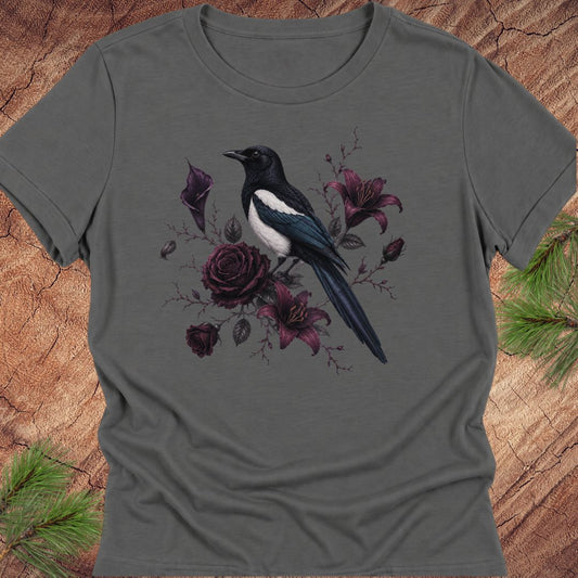 A dark gray short-sleeve crewneck t-shirt featuring a graphic print of a black and white magpie perched among dark purple roses and lilies, displayed flat on a rustic wood background with pine needles.